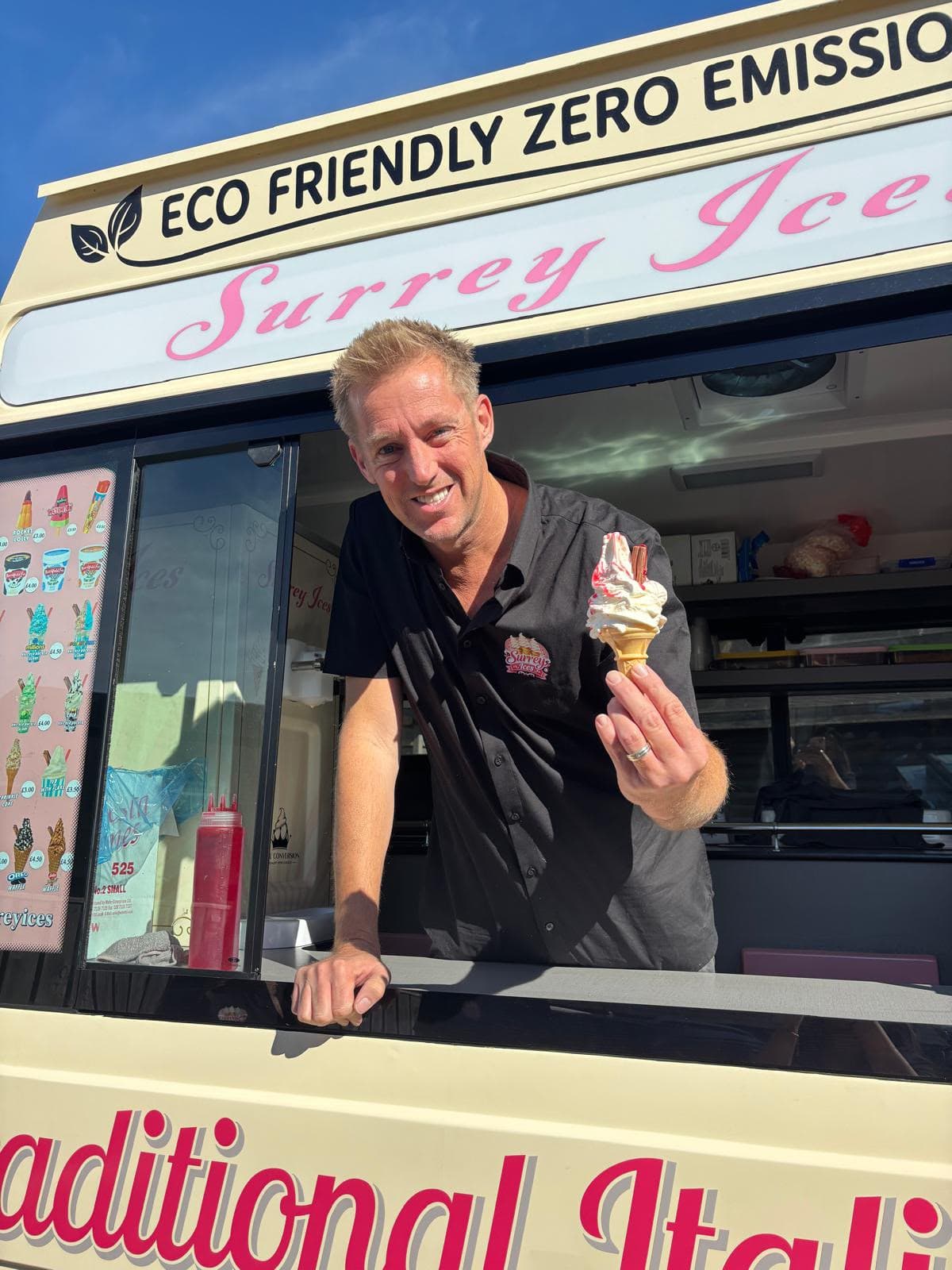 A Hampshire Ices server smiling in the van hatch and holding a soft serve cone with sauce and a flake