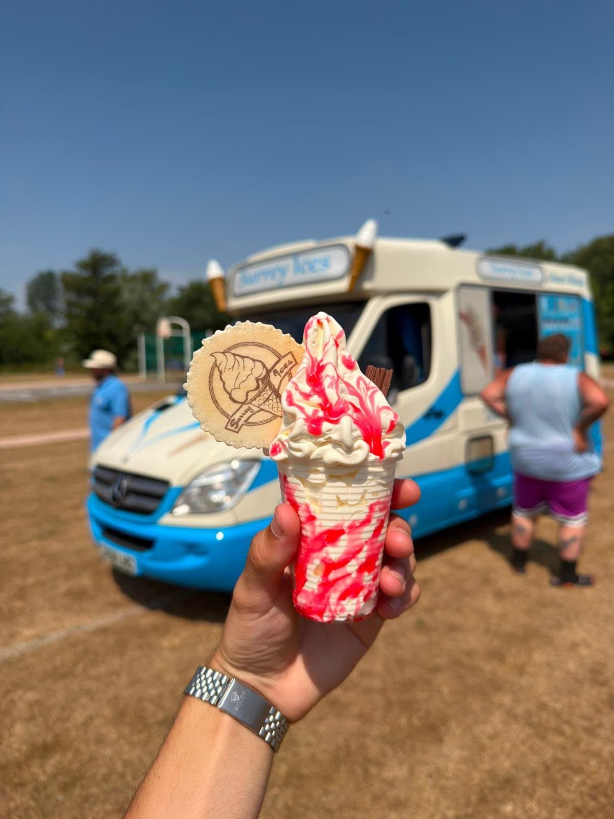 A hand holding a soft serve cone with red sauce and a Hampshire Ices wafer, with a blue and cream van on a playing field behind